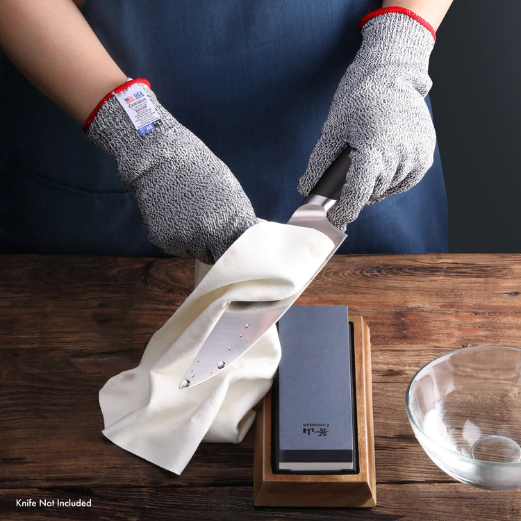 Hands wearing gray cut resistant gloves sharpening a knife on a whetstone with a wooden base.