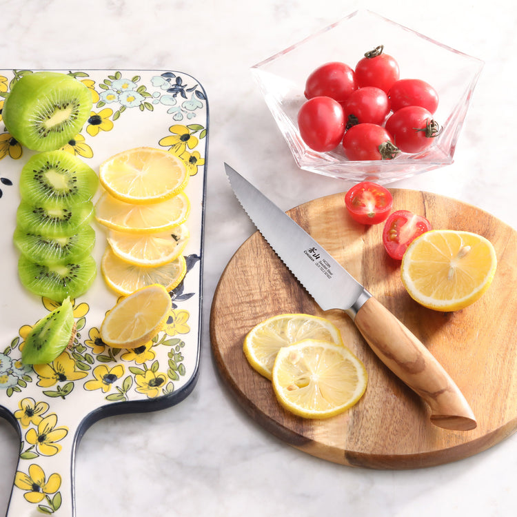 Serrated utility knife with olivewood handle on a round cutting board, sliced lemons and cherry tomatoes nearby.