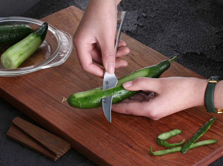Hands peeling a cucumber with a small curved knife on a wooden cutting board, with a wooden sheath nearby.