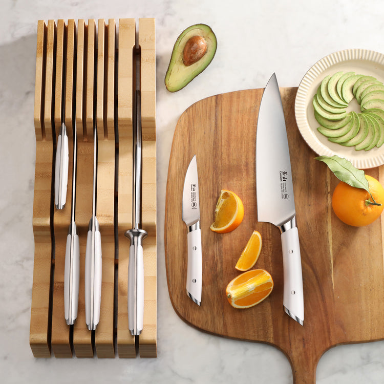 Seven-piece knife set with stainless steel blades and white handles in a bamboo in-drawer organizer, next to a wooden cutting