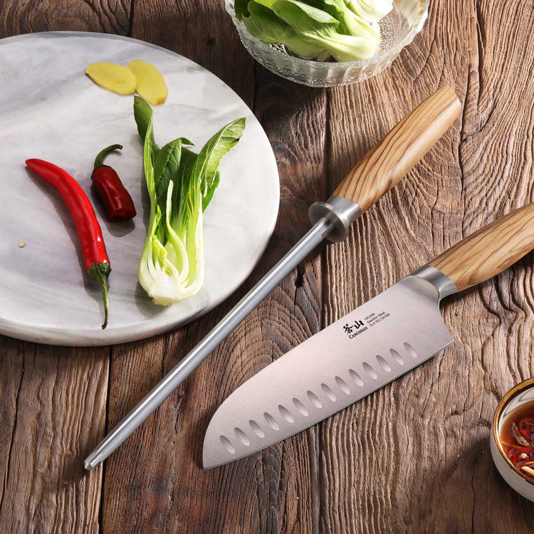 High-carbon honing steel with olivewood handle beside a chef's knife, bok choy, chili peppers, and ginger on a wooden surface