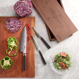
                    
                      Three kitchen knives with dark handles on a wooden board next to sliced vegetables and a wooden box with knife outlines.
                    
                  