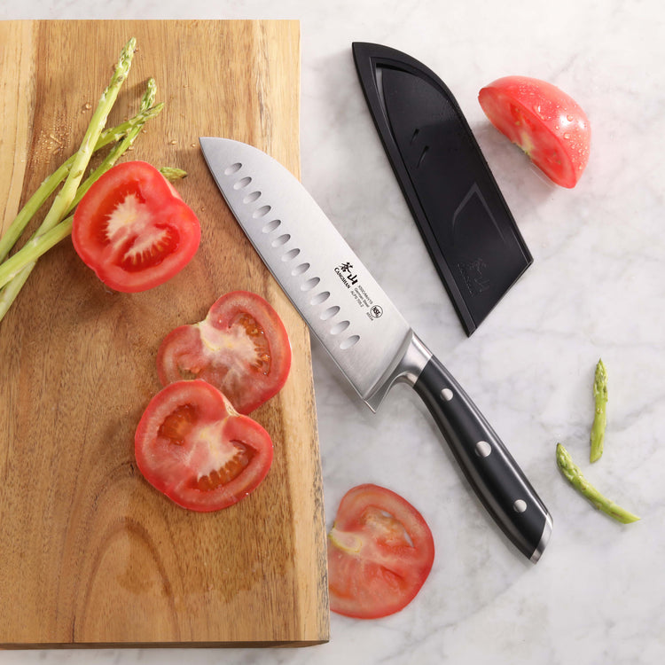 7-inch Santoku knife with black handle and sheath beside sliced tomato and asparagus on wooden board and marble surface.
