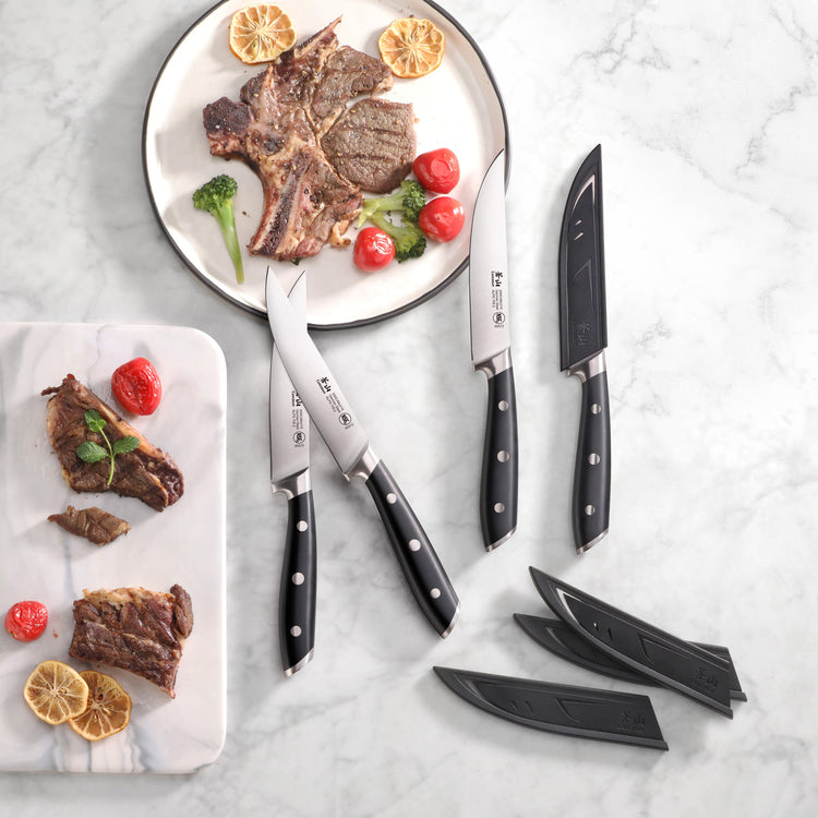 Four black-handled steak knives with sheaths next to cooked steak and vegetables on white plates on a marble surface.