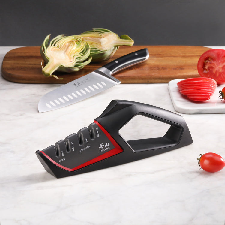Black and red handheld knife sharpener with coarse and fine slots on a marble countertop near a chef's knife, artichoke halve