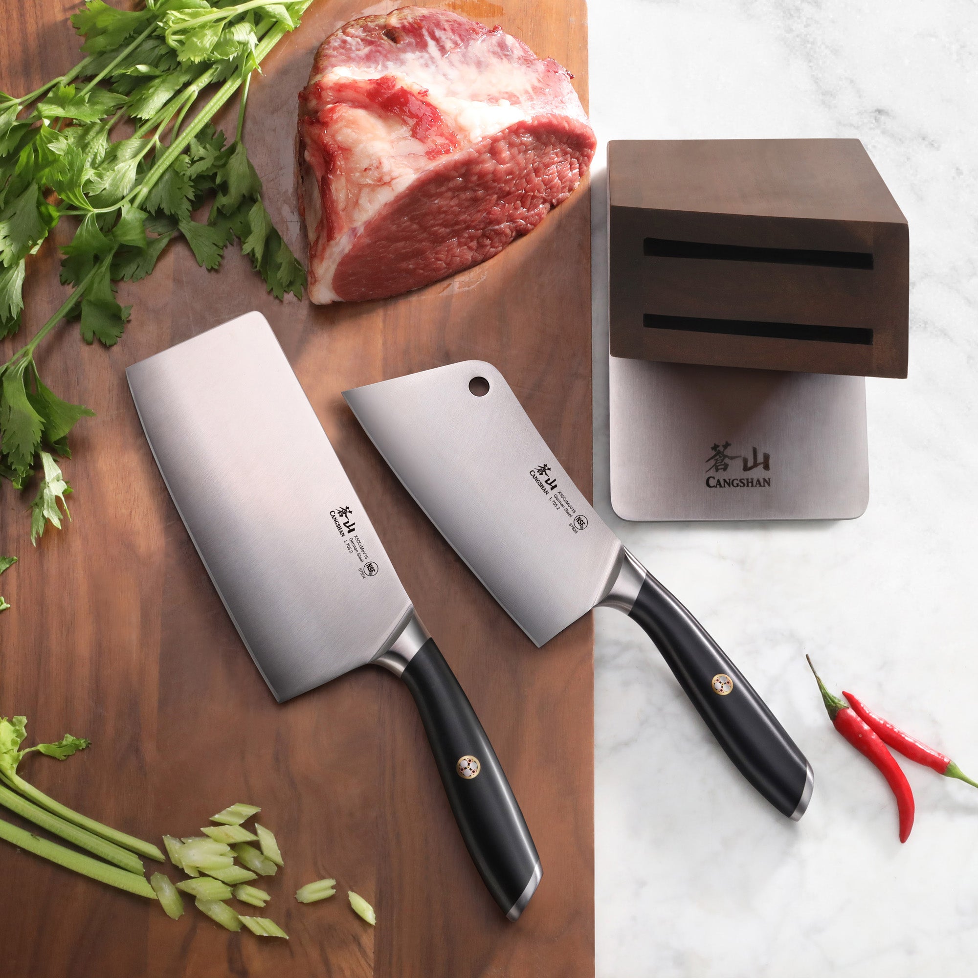 Two forged German steel cleavers with black handles next to a black acacia wood knife block, raw meat, and fresh herbs.