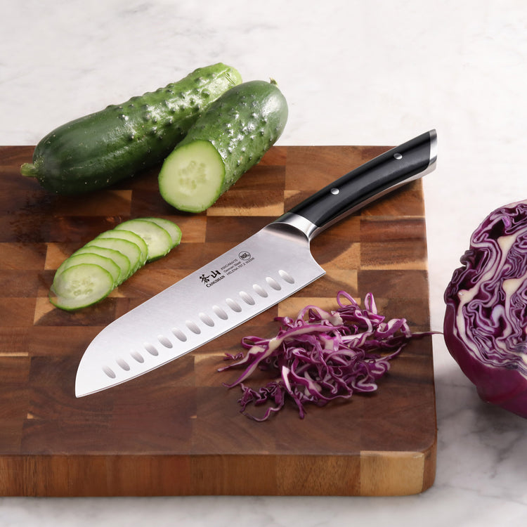 Santoku knife with black handle on wooden cutting board next to sliced cucumber and shredded purple cabbage.