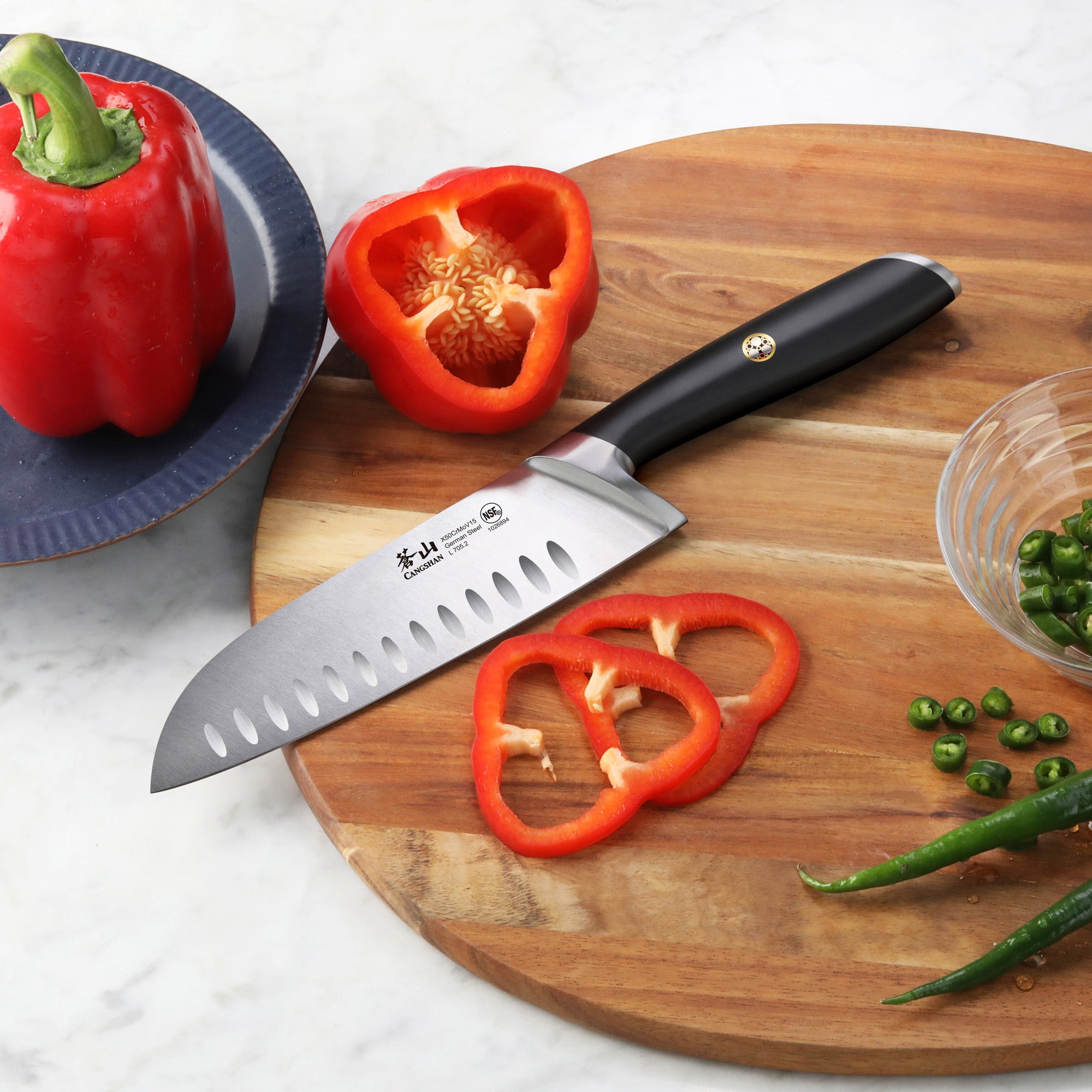 7-inch Santoku knife with black handle slicing red bell pepper on wooden cutting board with green chili peppers nearby.