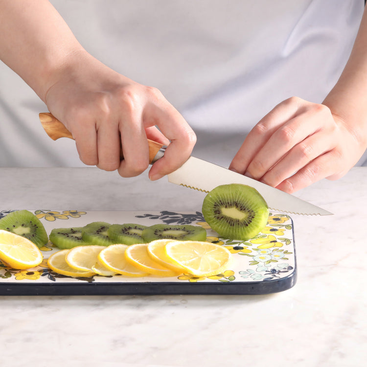 Hands holding a serrated knife with an olivewood handle, slicing a kiwi on a floral cutting board with lemon slices.