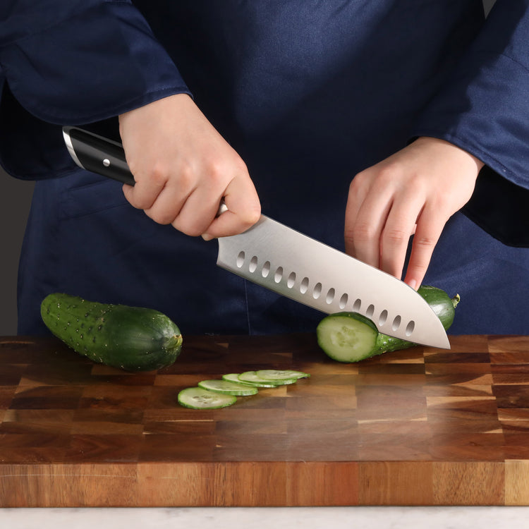 Hands slicing a cucumber into thin rounds on a wooden cutting board using a 7-inch Santoku knife with a black handle.