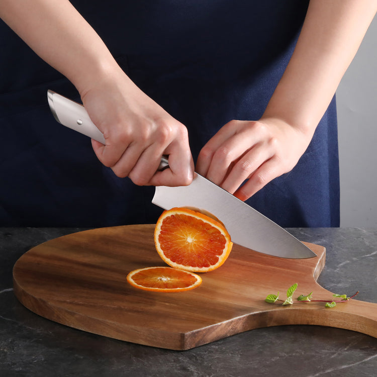 Hands slicing an orange with a white-handled knife on a wooden cutting board.