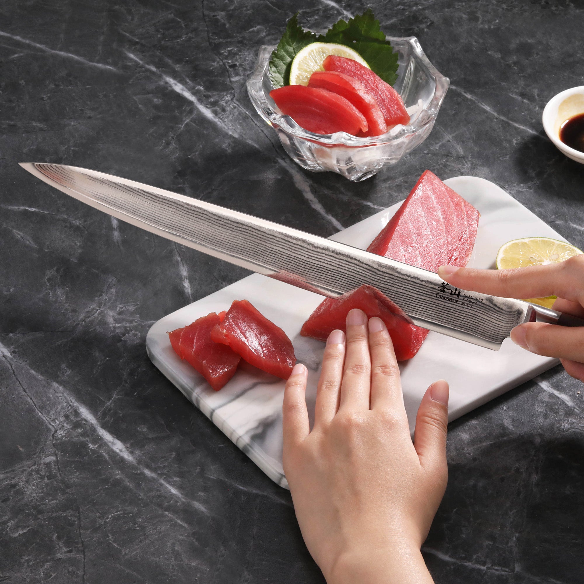 Hand holding a 12-inch Damascus steel sashimi knife slicing raw tuna on a white cutting board with a bowl of sashimi and lemo
