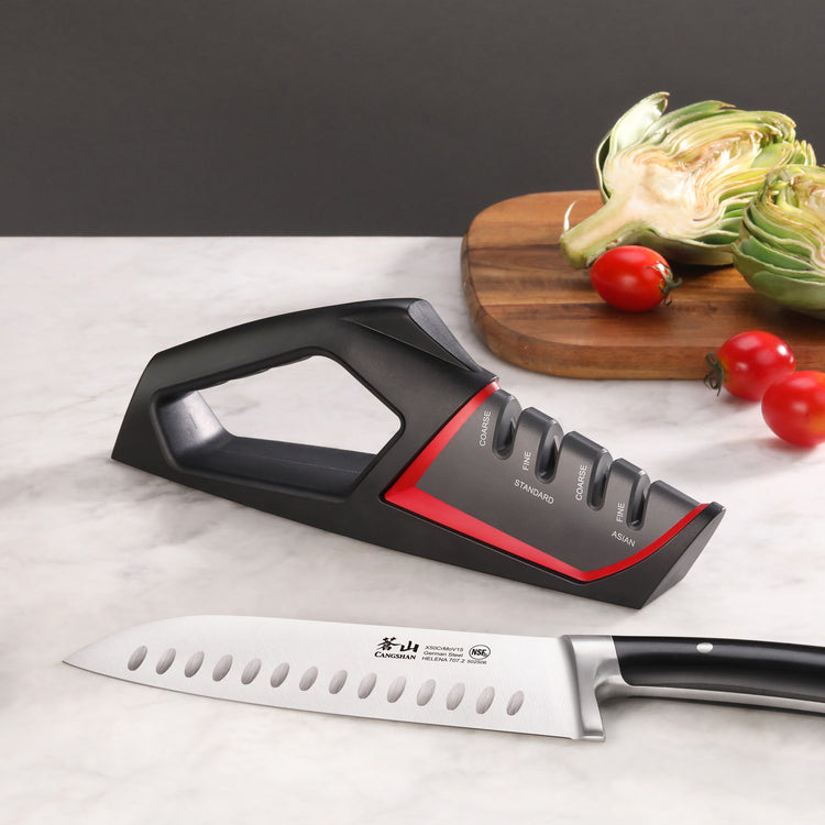 Black and red handheld knife sharpener with four sharpening slots placed on a marble surface next to a chef's knife and a cut