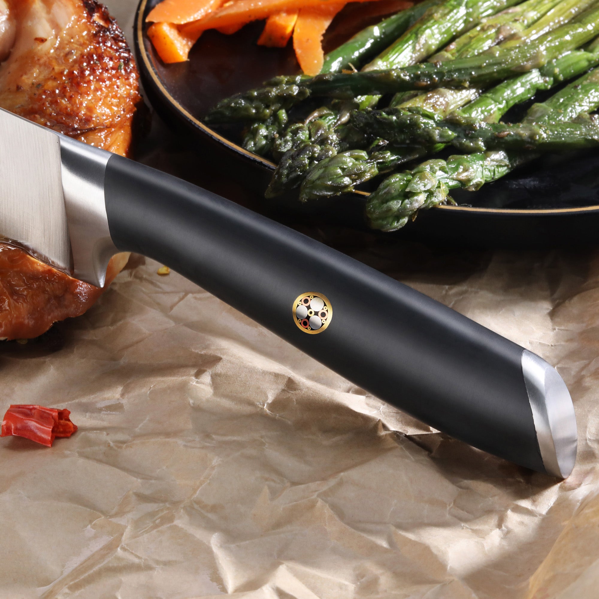 Black-handled carving knife resting on crumpled paper near roasted meat and a plate of asparagus and carrots.