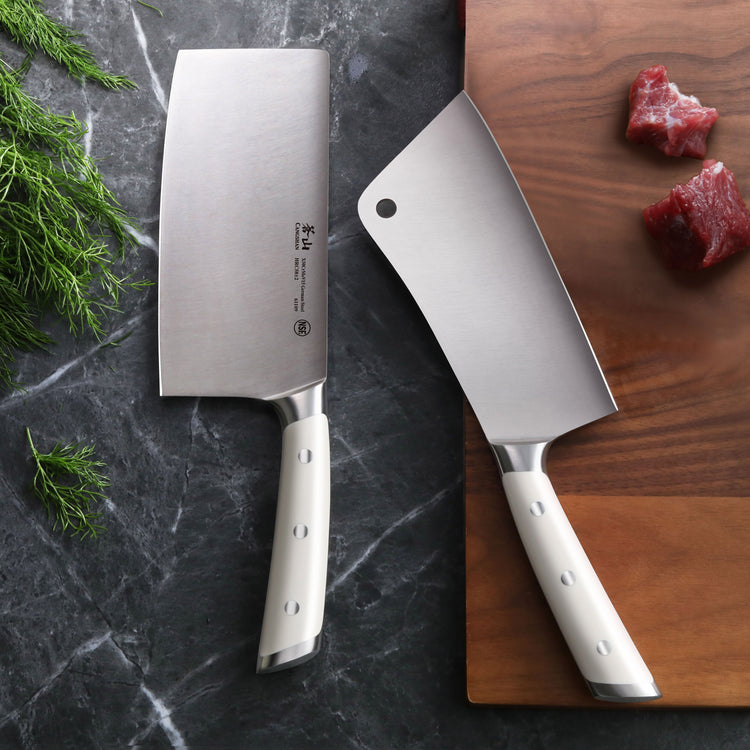 Two cleaver knives with white handles on a dark marble surface and wooden cutting board with pieces of raw meat.