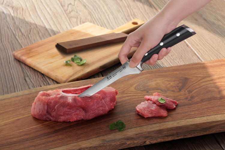 Hand holding a boning knife cutting raw meat on a wooden board, with a wooden sheath and herbs nearby.