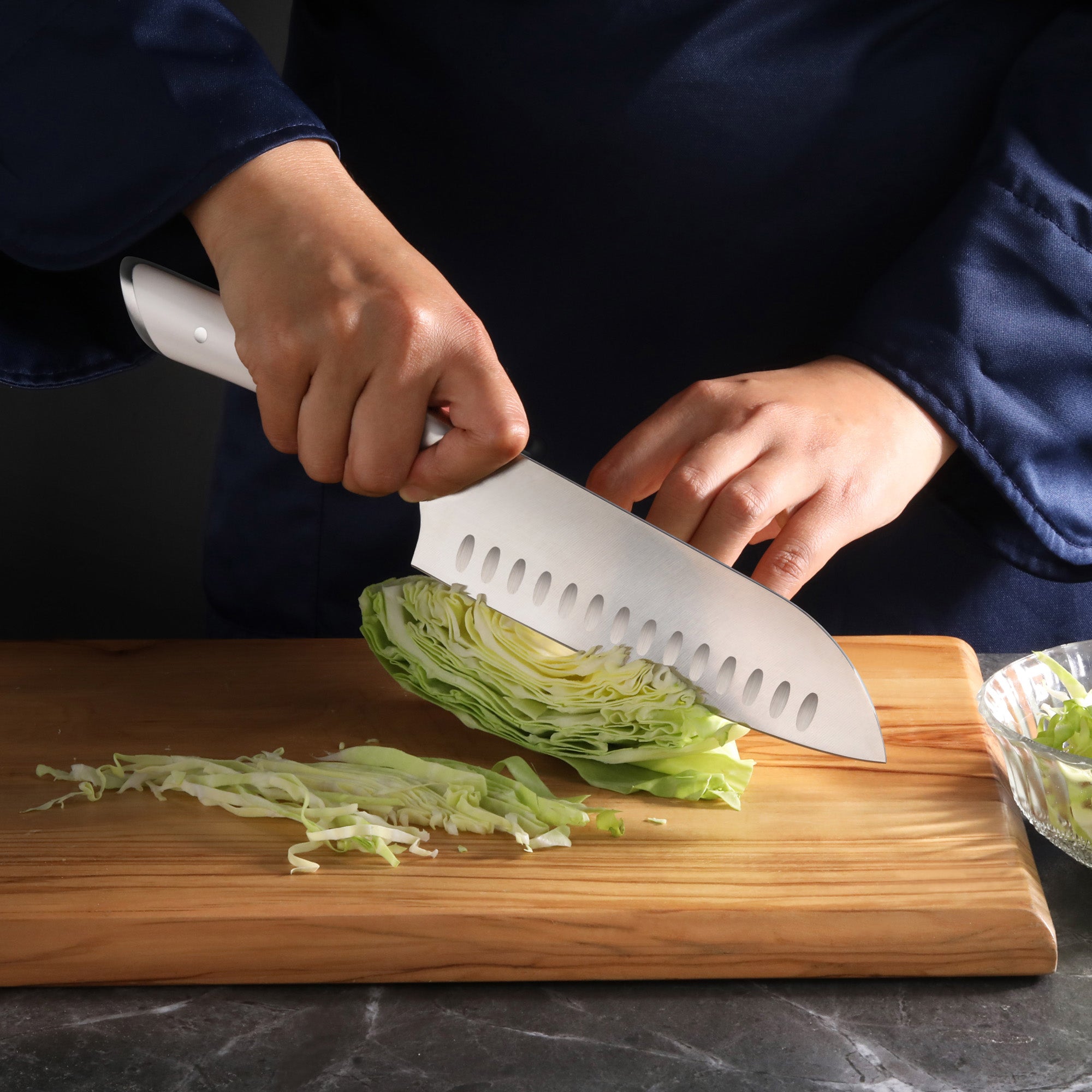 Hand holding a 7-inch Santoku knife slicing a wedge of cabbage on a wooden cutting board.