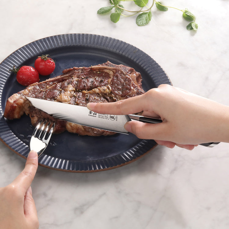 Hand holding a steak knife cutting a cooked steak on a blue plate with two cherry tomatoes and a fork holding the steak.