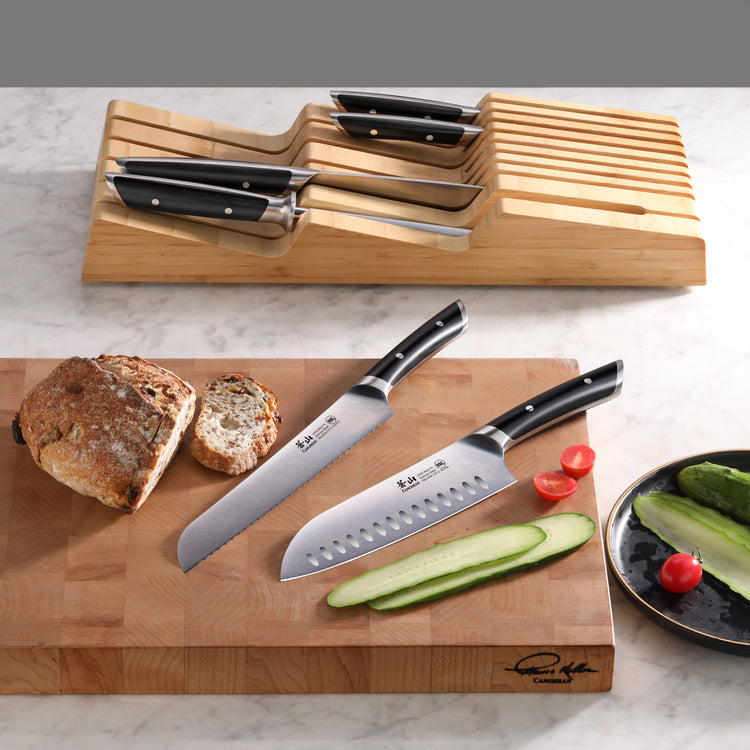 Seven-piece knife set with black handles in a bamboo in-drawer tray, two knives on a cutting board with bread and sliced cucu