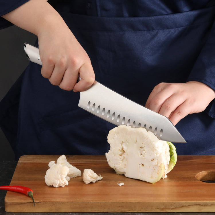 Hands using a large chef's knife to cut a cauliflower head on a wooden cutting board.