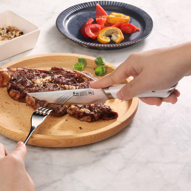 Hand using a steak knife and fork to cut a cooked steak on a wooden plate with grilled vegetables on a blue plate in the back