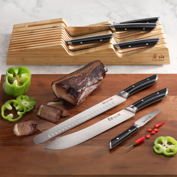 Wooden in-drawer knife organizer holding four knives with black handles, alongside three knives, sliced meat, green bell pepp