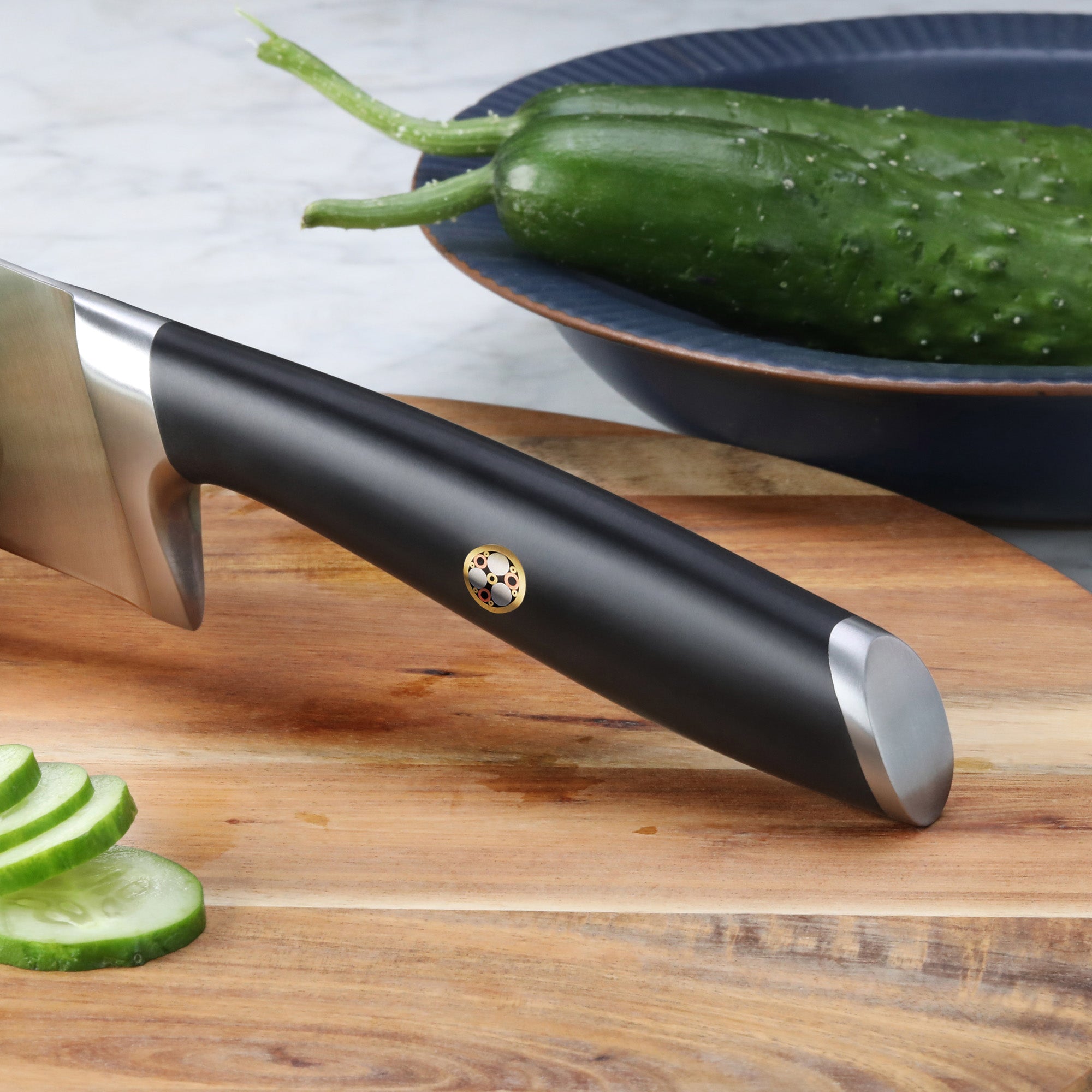 Black-handled Santoku knife on wooden cutting board with sliced cucumber and whole cucumbers in a blue bowl.