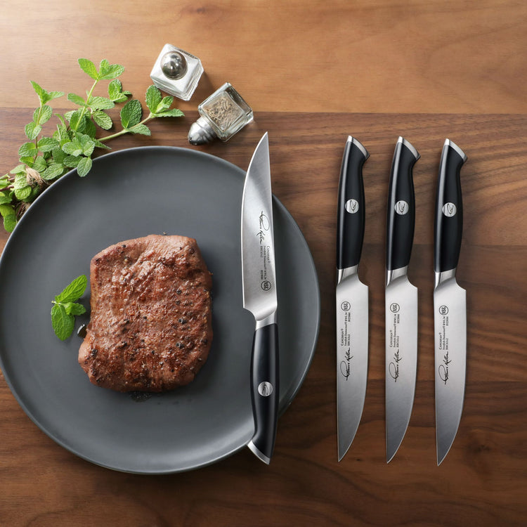 Four black-handled steak knives with stainless steel blades next to a cooked steak on a gray plate with herbs and salt and pe