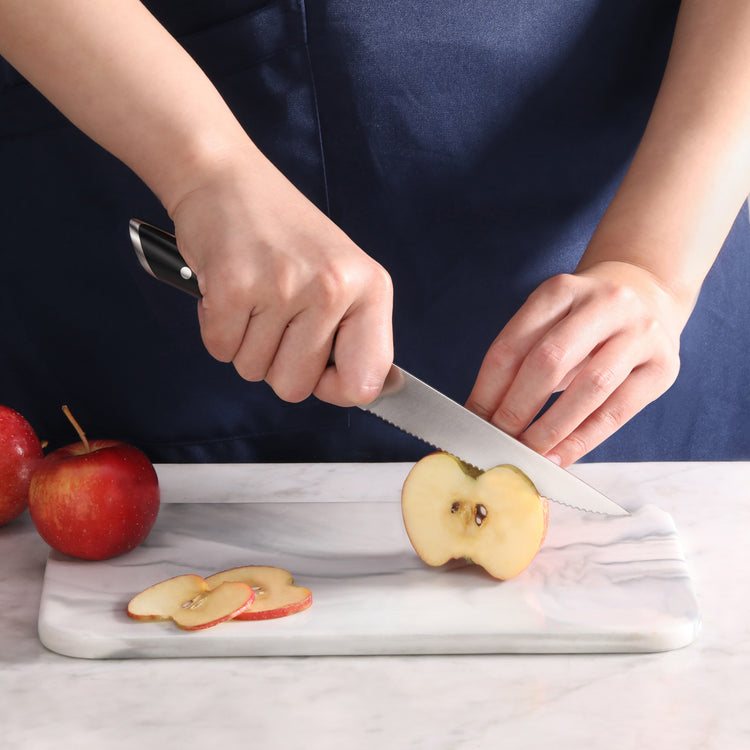 Hands holding a serrated utility knife slicing an apple on a marble cutting board with two whole apples nearby.