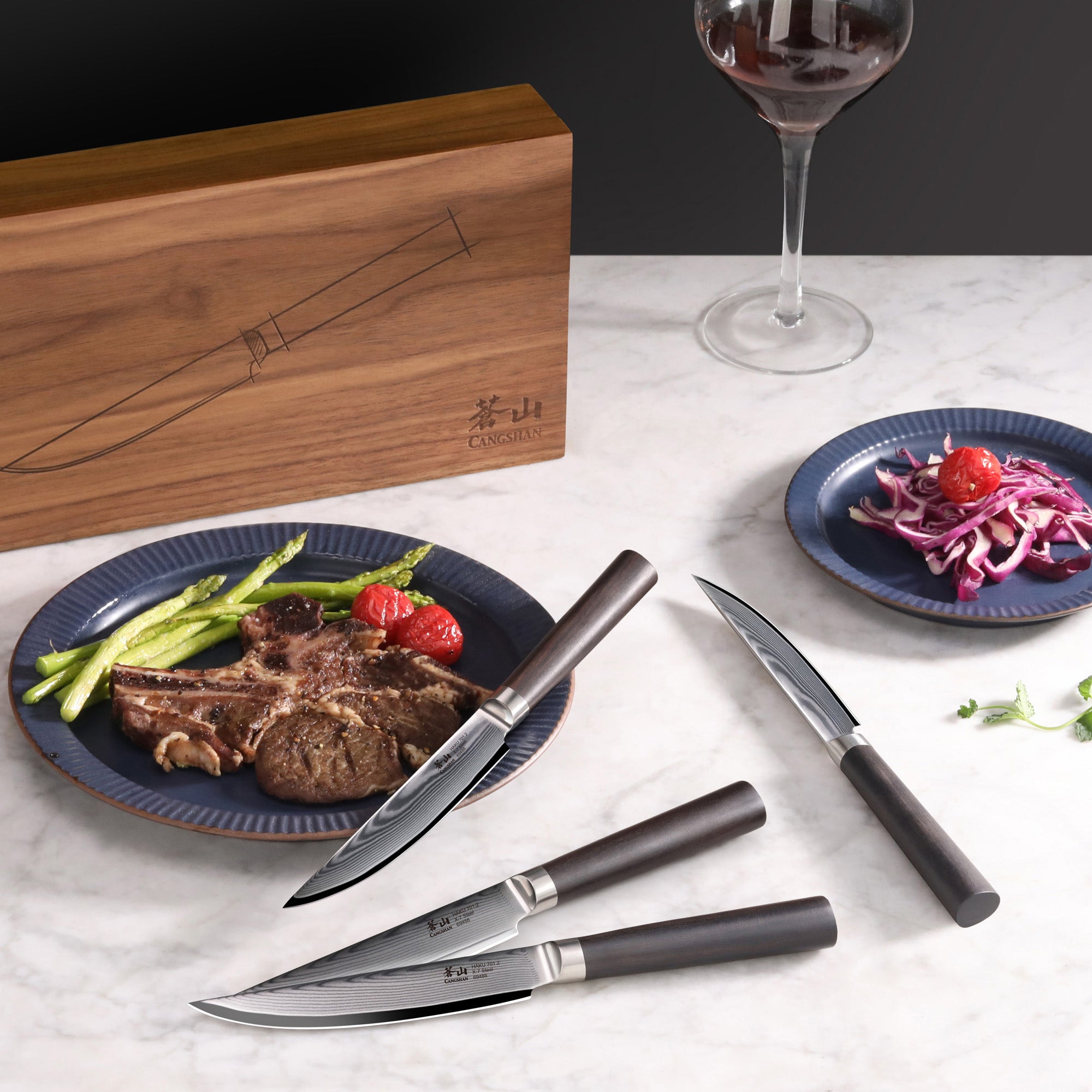 Four steak knives with dark handles and Damascus steel blades displayed on a marble surface, with a wooden box, plates of ste