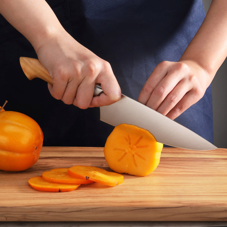 Hands slicing a persimmon on a wooden cutting board with a chef's knife featuring a wooden handle.