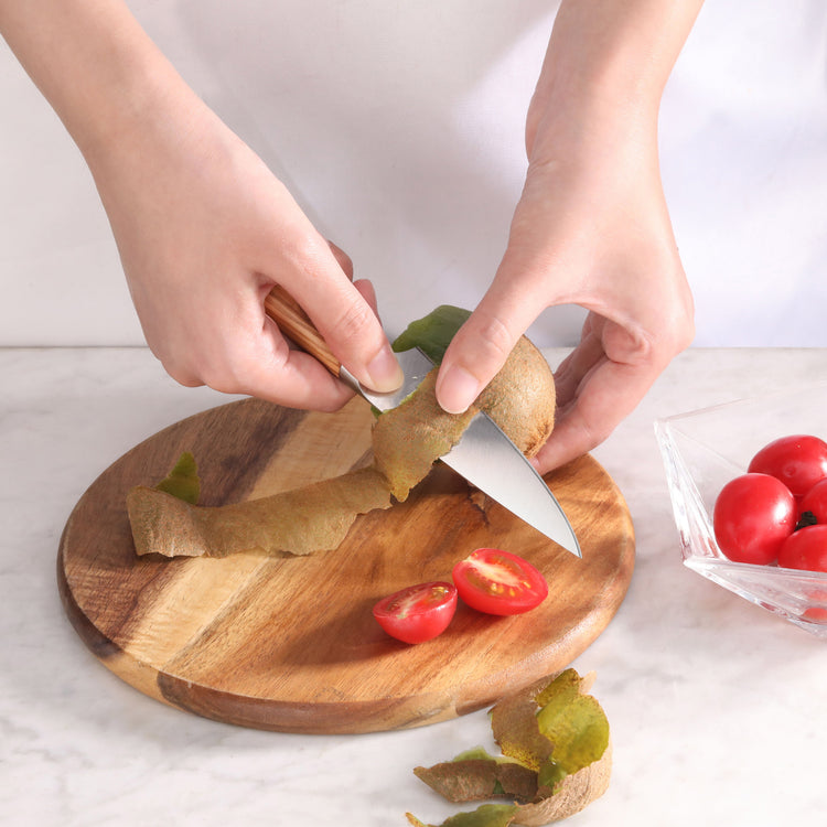 Hands peeling a kiwi with a small knife over a wooden cutting board next to halved cherry tomatoes.