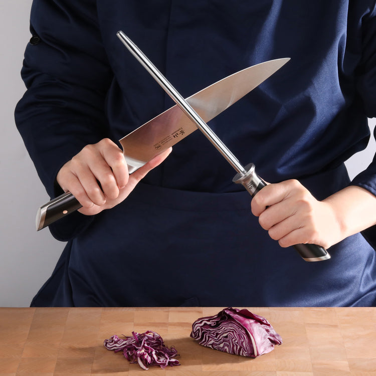 Person holding a knife and honing steel rod, sharpening the knife over a wooden surface with chopped purple cabbage.