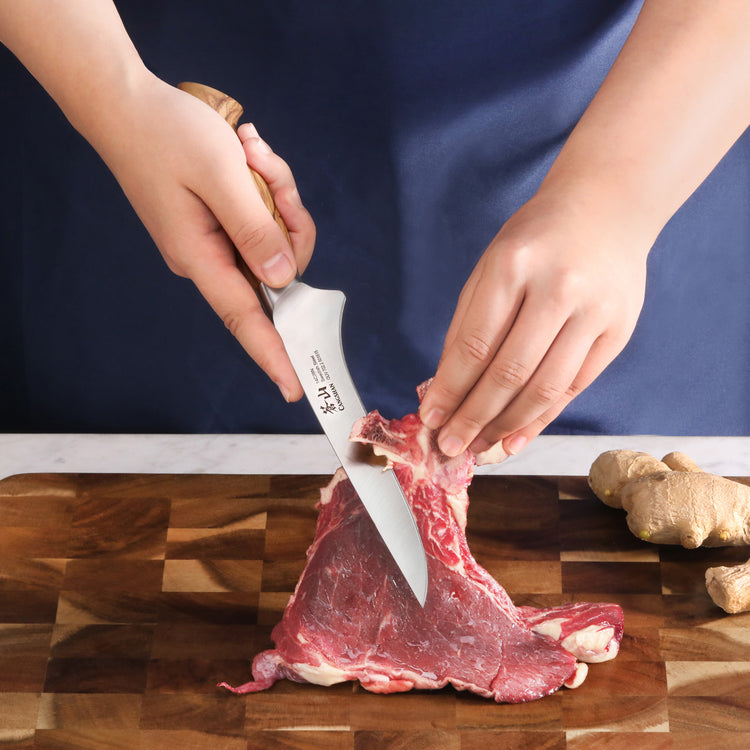 Hands holding a boning knife with an olivewood handle cutting raw meat on a wooden board.