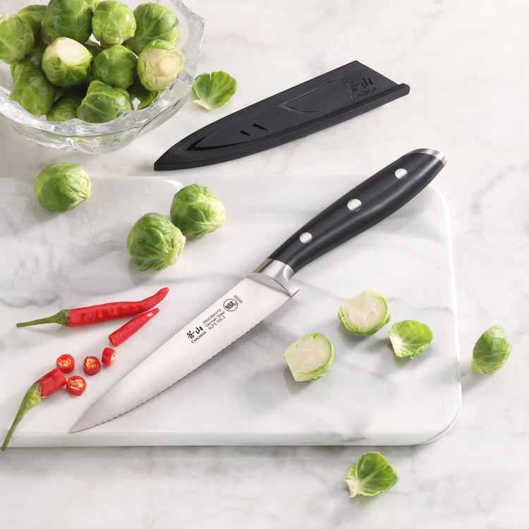 Serrated utility knife with black handle on white cutting board, surrounded by halved Brussels sprouts and red chili peppers,