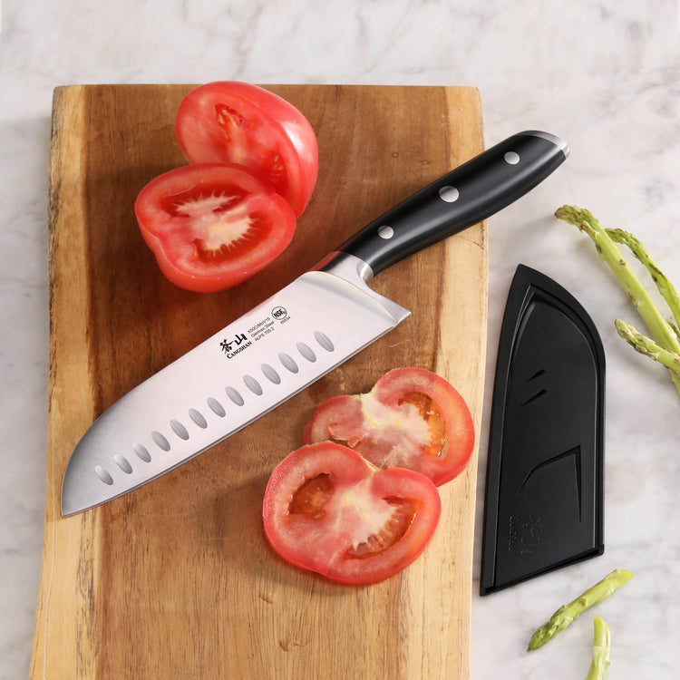 Santoku knife with black handle on wooden cutting board next to sliced tomato and black sheath.