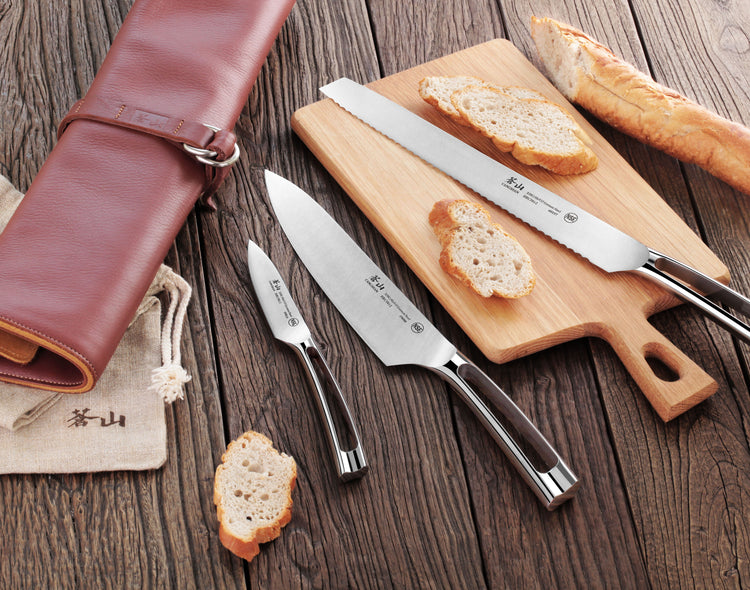 Four stainless steel knives with black handles displayed on a wooden surface with sliced bread and a brown leather knife roll