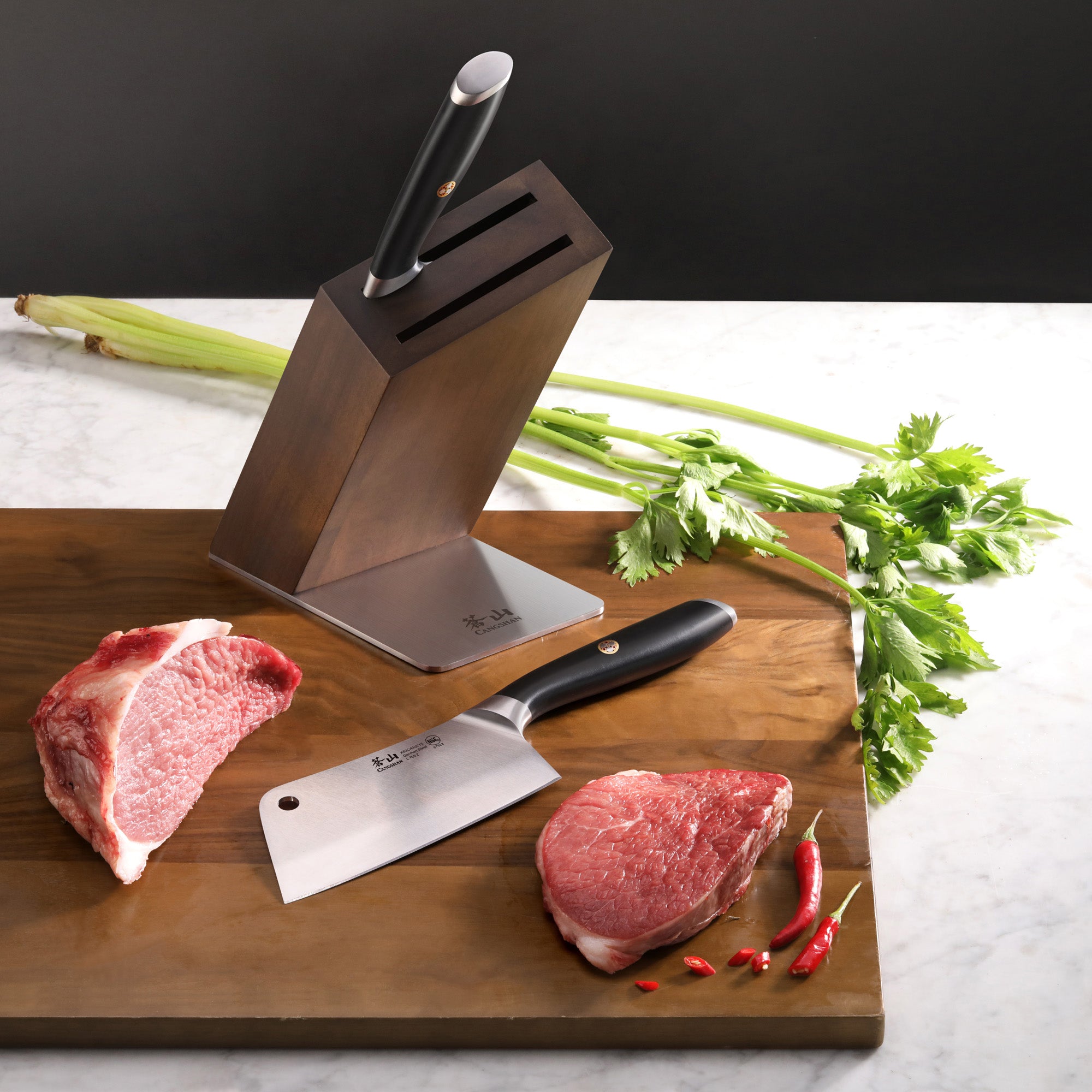 Wooden knife block with two cleaver knives, raw beef cuts, celery stalks, and red chili peppers on a marble surface.
