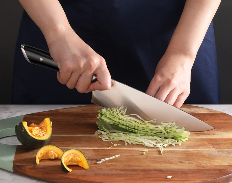 Hands slicing shredded cabbage on a wooden cutting board with a large kitchen knife and pumpkin pieces nearby.