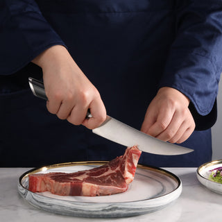 
                    
                      Person holding a boning knife cutting a raw steak on a white plate with a gold rim.
                    
                  