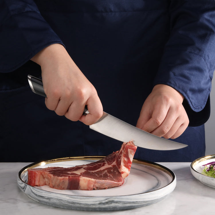 Person holding a boning knife cutting a raw steak on a white plate with a gold rim.