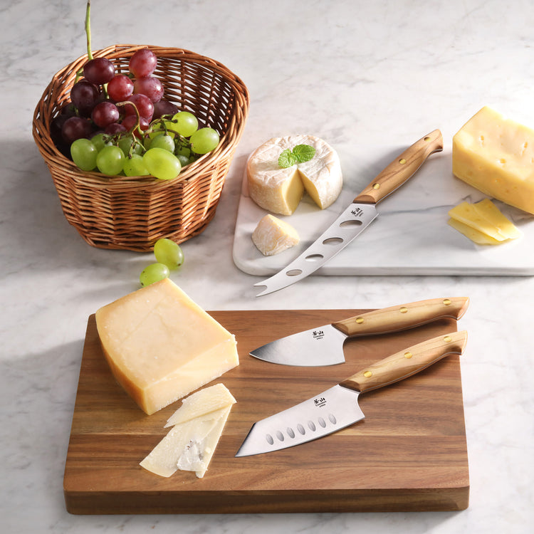 Three olive wood-handled cheese knives on an acacia wood board with sliced cheese, a basket of grapes, and a round cheese whe