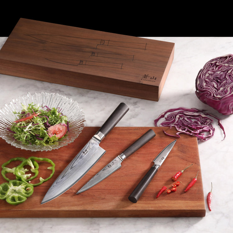 Three Damascus steel knives with dark handles on a wooden cutting board, surrounded by sliced vegetables and a wooden box.