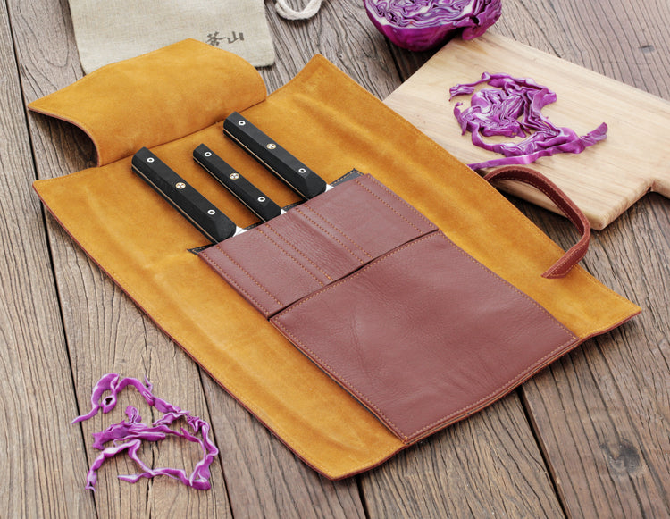 Four knives with black handles in a brown leather roll set on a wooden surface with sliced purple cabbage.