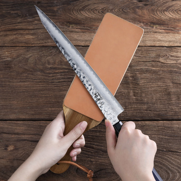 Hands holding a leather paddle strop and sharpening a large hammered steel knife blade on a wooden surface.