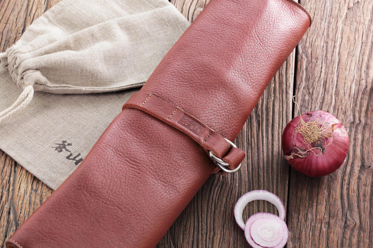 Rolled brown leather knife case secured with a strap, next to a beige drawstring pouch and a whole and sliced red onion on a