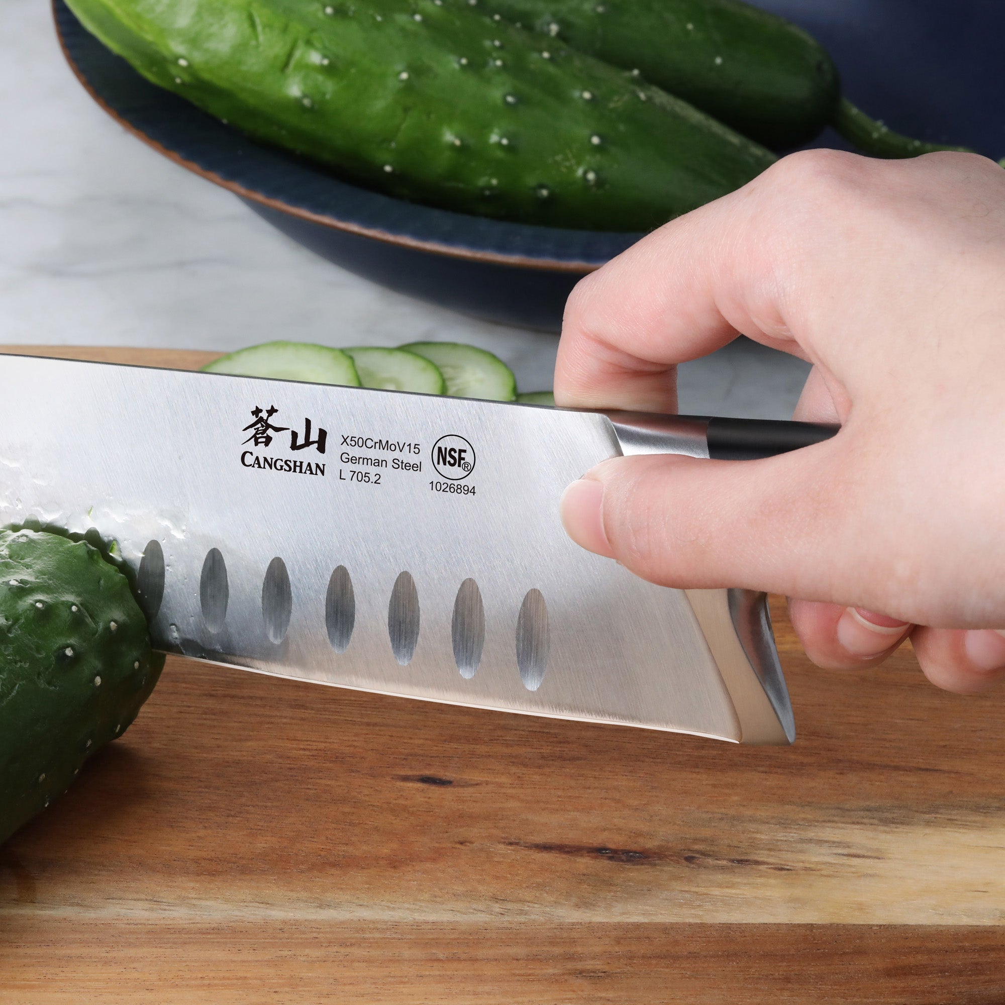 Hand holding a 7-inch Santoku knife slicing a cucumber on a wooden cutting board with whole cucumbers in the background.