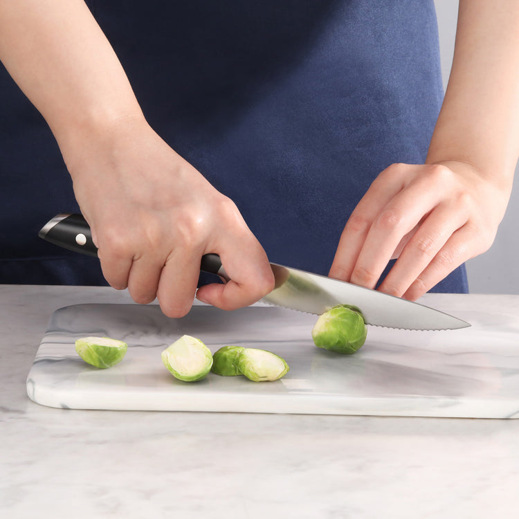 Hands slicing Brussels sprouts on a marble cutting board with a serrated utility knife with black handle.