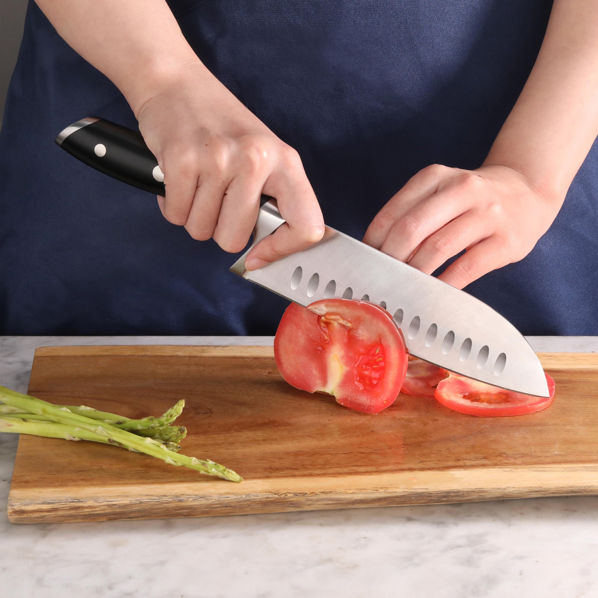 Hands slicing a tomato on a wooden cutting board with a black-handled Santoku knife, asparagus nearby.