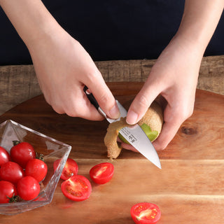 
                    
                      Hands peeling a kiwi with a small black-handled paring knife on a wooden cutting board next to halved cherry tomatoes.
                    
                  